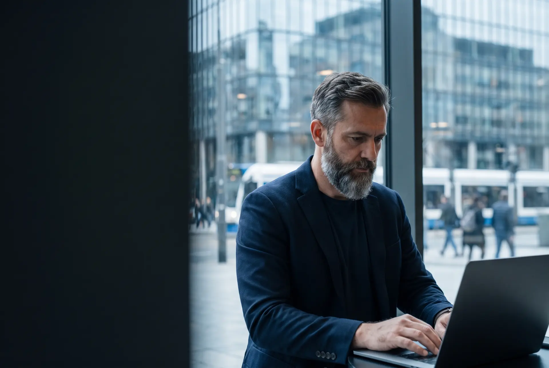 Man with a grey beard working on a laptop in front of a glass office window